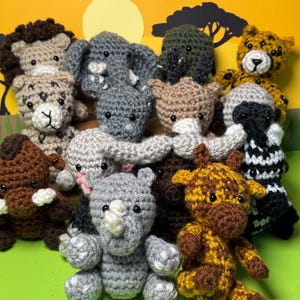Puede incluir: Una colección de juguetes de animales de safari hechos a mano con ganchillo, que incluyen un león, un elefante, un guepardo, un rinoceronte, una jirafa y una cebra. Los juguetes son de varios colores, con detalles y una textura suave. El fondo es una escena colorida de una puesta de sol.