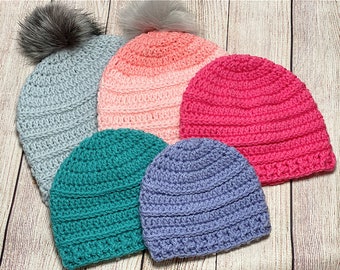 May include: Five crocheted hats in different colors, including gray, pink, teal, and purple. Each hat has a ribbed texture and a pompom on top.
