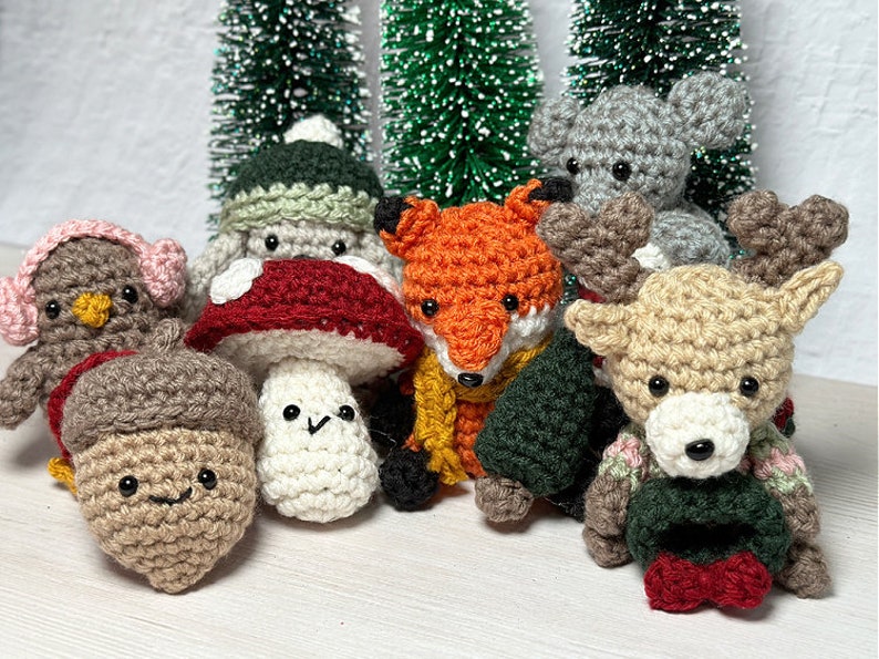 K&ouml;nnte beinhalten: Eine Sammlung geh&auml;kelter Waldtiere, darunter ein Pilz, eine Eichel, ein Fuchs, ein Rentier und ein graues Kaninchen. Die Tiere tragen alle winterliche Kleidung und sind vor einem Hintergrund aus Miniatur-Weihnachtsb&auml;umen angeordnet.