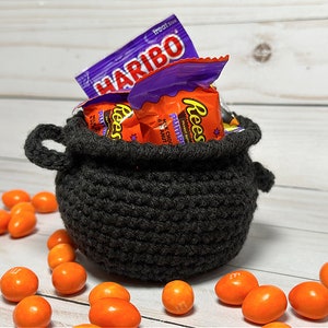 May include: A black crocheted cauldron filled with Halloween sweets, including Reese's Peanut Butter Cups and Haribo gummy sweets. Orange and yellow sweets are scattered around the cauldron on a white wooden surface.