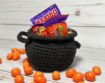 Op de afbeelding: Een zwarte gehaakte ketel gevuld met Halloween-snoep, waaronder Reese's Peanut Butter Cups en Haribo gummy snoepjes. Oranje en gele snoepjes zijn rond de ketel verspreid op een wit houten oppervlak.