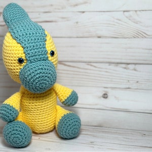 May include: A crocheted dinosaur toy with a yellow body and blue head and feet. The dinosaur has large blue eyes and a small, pointed nose.