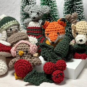 K&ouml;nnte beinhalten: Eine Sammlung geh&auml;kelter Weihnachtsornamente, darunter ein Fuchs, ein Rentier, ein Pilz, ein Vogel und ein Schneemann. Die Ornamente sind mit Stechpalmen und Beeren verziert.