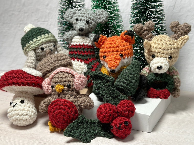K&ouml;nnte beinhalten: Eine Sammlung geh&auml;kelter Weihnachtsornamente, darunter ein Fuchs, ein Rentier, ein Pilz, ein Vogel und ein Schneemann. Die Ornamente sind mit Stechpalmen und Beeren verziert.