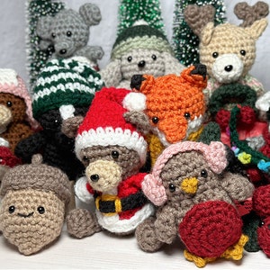 K&ouml;nnte beinhalten: Eine Sammlung von geh&auml;kelten Weihnachtsornamenten, darunter ein Fuchs, ein Rentier, ein B&auml;r mit einem Weihnachtsmann-Hut, ein Pilz, eine Eichel, ein Vogel und ein Waschb&auml;r. Die Ornamente sind alle aus verschiedenen Garnfarben gefertigt und haben unterschiedliche Gesichtsausdr&uuml;cke.