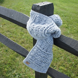 May include: A light blue crocheted scarf draped over a black wooden fence post. The scarf has a textured, cable knit pattern.