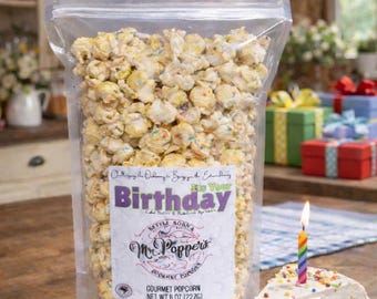 May include: A clear plastic bag of Mr. Popper's gourmet popcorn with a label reading It's Your Birthday, Cake Batter & Rainbow Sprinkles, Net Wt 8 oz (227g). Next to it is a slice of cake with white frosting, rainbow sprinkles, and a lit rainbow candle.