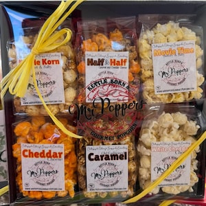 May include: A gift box of gourmet popcorn in clear bags, tied with a yellow ribbon. Flavors include kettle corn, cheddar cheese, caramel, and movie time. The brand name "Mr. Poppers" is visible on the labels.