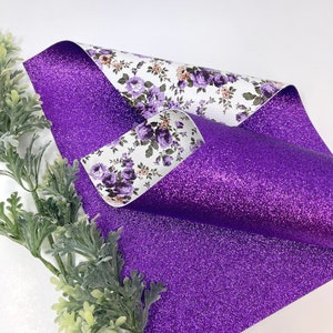 May include: A sheet of purple glitter fabric is curled to reveal a floral pattern on the reverse side. The floral pattern features purple and tan flowers on a white background. Green leafy sprigs are in the lower left corner.