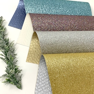 May include: An assortment of glitter and textured craft paper in shades of blue, purple, silver, gold, and white. The papers are arranged in a layered display, showcasing their varying textures and finishes. A sprig of greenery is in the corner.