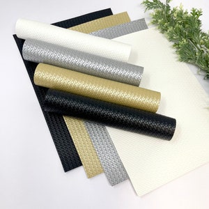 May include: Assortment of textured, woven-look vinyl rolls and sheets in various colors, including black, gold, silver, and white. The rolls are partially unrolled, revealing the material's pattern. A sprig of greenery is in the background.