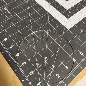 Puede incluir: Una regla circular de pl&aacute;stico transparente con marcas para grados y pulgadas. La regla est&aacute; sobre una estera de corte gris con l&iacute;neas blancas.