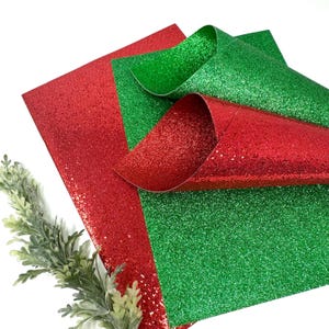 Puede incluir: Hojas de purpurina roja y verde dispuestas en una composición en capas. La hoja roja está plana, mientras que la hoja verde está parcialmente enrollada. La purpurina refleja la luz, creando un efecto brillante. Una ramita de follaje verde está en la esquina inferior izquierda.