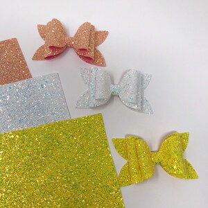 May include: Three glitter fabric sheets in pink, white, and yellow. Each sheet has a different glitter pattern. Three glitter bows are also shown, one in each color.