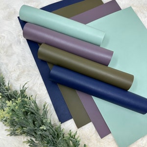 May include: An assortment of colorful faux leather sheets and rolls in shades of blue, green, purple, and olive. The sheets are flat, while some are rolled up. The background is a soft, white, textured surface.