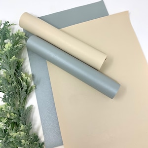 Puede incluir: Dos rollos de piel sintética en beige claro y azul claro. Los rollos están dispuestos sobre una superficie blanca.