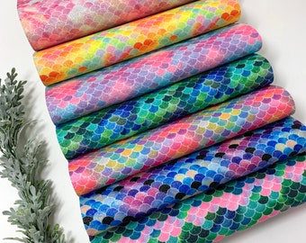 May include: A stack of colorful faux leather sheets with a mermaid scale pattern. The colors include pink, rainbow, purple, green, and blue. The sheets are stacked on top of each other, with a sprig of greenery on the left side.