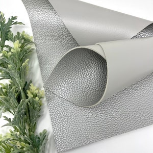 May include: A sheet of silver faux leather with a textured surface. The leather is folded to show the smooth, light gray underside.