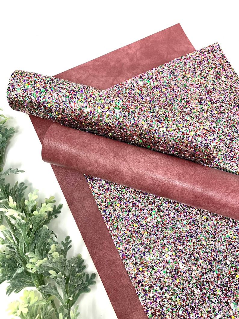 May include: A sheet of burgundy faux leather with a sheet of multi-colored glitter fabric on top. The glitter fabric has a white base with red, green, blue, and purple glitter.