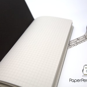 May include: Open notebook with grid paper pages and a black cover. A roll of decorative tape with text is next to the notebook. The tape is gray with black text. The notebook is on a white surface.
