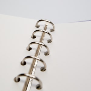 May include: Close-up of an open notebook with a silver metal binder. The notebook pages are white, and the binder rings are curved and shiny. The background is a soft, neutral color.