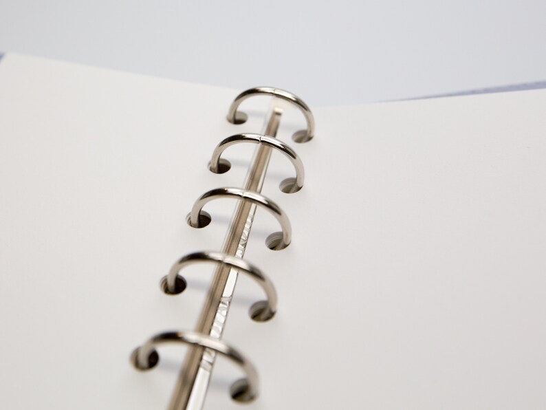 May include: Close-up of an open notebook with a silver metal binder. The notebook pages are white, and the binder rings are curved and shiny. The background is a soft, neutral color.