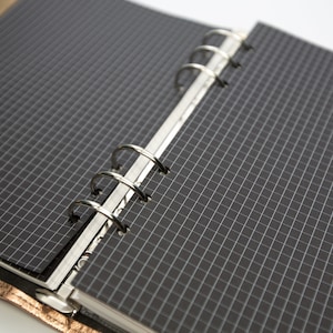 May include: Open planner with black grid paper and silver metal rings. The planner is open to reveal the interior pages, which are lined with a white grid pattern. The cover is a metallic gold color.