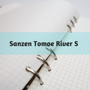 May include: Open notebook with grid paper. The notebook is open to a page with a light blue banner across the middle. The text "Sanzen Tomoe River S" is printed on the banner. The notebook has silver metal rings.