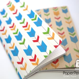 May include: Two notebooks with arrow patterns. One notebook has a white background with blue, green, red, and orange arrows. The other notebook has a tan background with the same arrow design. The notebooks are open, revealing lined pages.