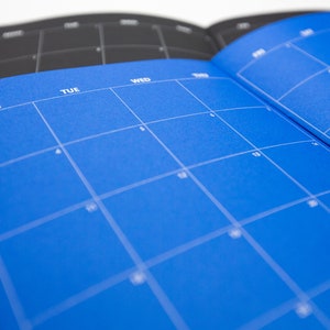 May include: Open planner with a bright blue page featuring a calendar grid. The days of the week are printed in white. A second planner with a black page is visible in the background. The planner is open to a monthly view.