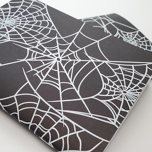 May include: Two black notebooks with white spiderweb designs. The spiderwebs are centered and cover the entire front of the notebooks. The notebooks are stacked on top of each other, with the top notebook slightly angled.