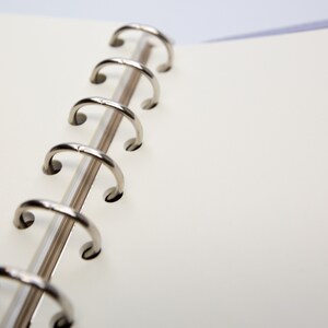 May include: Close-up of an open notebook with a silver metal binder. The notebook has blank, off-white pages. The binder is made of six curved metal rings. The background is a soft, blurred white.