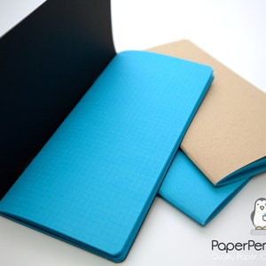 May include: An open black notebook next to two notebooks with blue and tan covers. The blue notebook is open, revealing the pages. The Paper Penguin Co. logo is in the bottom right corner.