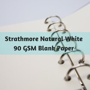 May include: Close-up of Strathmore Natural White 90 GSM blank paper in a binder. The paper is a bright white, and the binder rings are silver. The text is displayed on a light blue background.