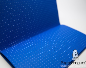 Peut inclure: Un cahier ouvert avec une couverture bleu vif ornée de petits points blancs. Le cahier est ouvert, révélant les pages intérieures. Le logo Paper Penguin Co. est visible en bas à droite.