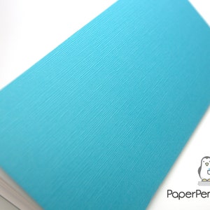 May include: A turquoise notebook with a textured cover is shown at an angle, revealing the white pages within. The cover has a subtle, diagonal ribbed pattern. The PaperPenguinCo logo is in the bottom right corner.