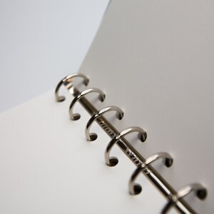 May include: Close-up of an open notebook with white pages and a silver metal binder. The binder has a series of curved rings that hold the pages together. The notebook is on a white background.