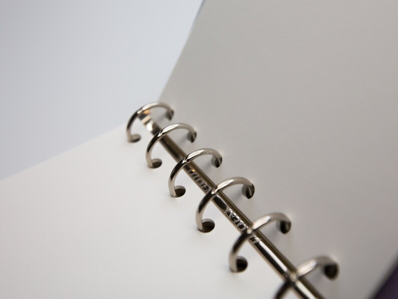 May include: Close-up of an open notebook with white pages and a silver metal binder. The binder has a series of curved rings that hold the pages together. The notebook is on a white background.