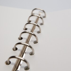 May include: Close-up of a silver metal binder mechanism with six open rings. The rings are attached to a silver metal spine. White paper pages are visible on the right side of the image.