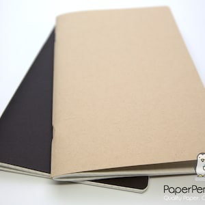 Puede incluir: Dos cuadernos abiertos, uno negro y otro beige, se muestran sobre una superficie blanca. El cuaderno beige est&aacute; encima, revelando sus p&aacute;ginas abiertas. El cuaderno negro es parcialmente visible debajo. El logotipo de PaperPenguinCo est&aacute; en la esquina inferior derecha.