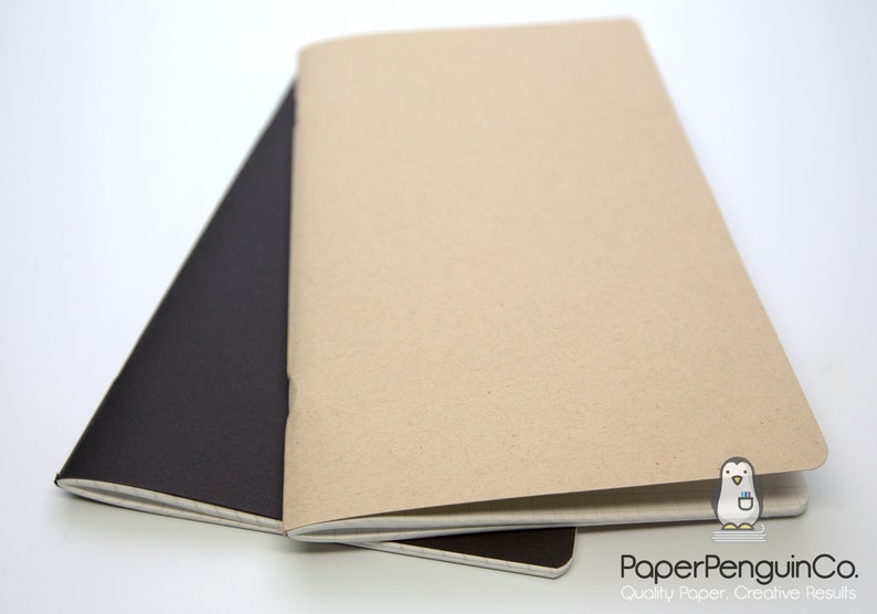 Puede incluir: Dos cuadernos abiertos, uno negro y otro beige, se muestran sobre una superficie blanca. El cuaderno beige est&aacute; encima, revelando sus p&aacute;ginas abiertas. El cuaderno negro es parcialmente visible debajo. El logotipo de PaperPenguinCo est&aacute; en la esquina inferior derecha.