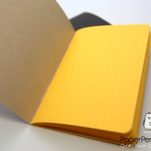 May include: Open notebook with a bright yellow dotted interior and a brown cover. The notebook is open to reveal the yellow pages, with a few pages turned. The PaperPenguinCo logo is in the bottom right corner.