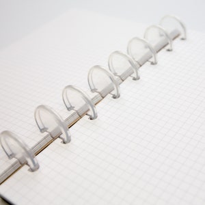 May include: Open notebook with white graph paper and clear plastic binding rings. The notebook is open, revealing the grid-lined pages. The binding rings are evenly spaced along the spine, holding the pages together.