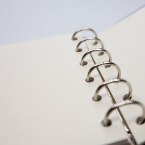 May include: Open notebook with blank, off-white pages. The silver metal rings of the binder are visible on the right side. The pages are slightly angled, suggesting the notebook is open. The background is a soft, blurred white and purple.