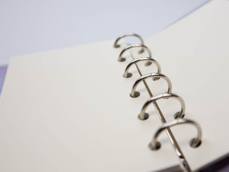 May include: Open notebook with blank, off-white pages. The silver metal rings of the binder are visible on the right side. The pages are slightly angled, suggesting the notebook is open. The background is a soft, blurred white and purple.