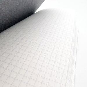Puede incluir: Cuaderno abierto con papel cuadriculado blanco y una cubierta gris oscuro. Las p&aacute;ginas est&aacute;n ligeramente curvadas, revelando el patr&oacute;n de cuadr&iacute;cula. El cuaderno est&aacute; sobre un fondo blanco, creando una est&eacute;tica limpia y minimalista.