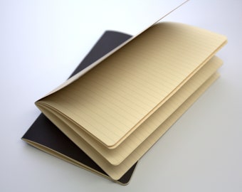 Può includere: Due quaderni, uno aperto, con pagine a righe color crema. Il quaderno inferiore è chiuso e nero. Le pagine del quaderno aperto sono leggermente curve, rivelando l'interno. Lo sfondo è bianco.