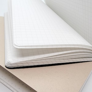 Puede incluir: Dos cuadernos abiertos. Uno est&aacute; abierto, revelando p&aacute;ginas cuadriculadas blancas. El otro est&aacute; cerrado, con una cubierta marr&oacute;n claro. Las p&aacute;ginas son blancas y la encuadernaci&oacute;n es negra.