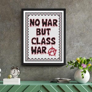 Könnte beinhalten: Ein gerahmtes Kreuzstich-Kunstwerk mit den Worten "NO WAR BUT CLASS WAR" in roten, tropfenden Buchstaben. Anarchie-Symbol unten. Das Kunstwerk hängt an einer grauen Wand über einem grünen Schrank.