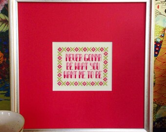 May include: A framed cross-stitch with a white background and a pink and green border. The cross-stitch says "Never gonna be what you want me to be."